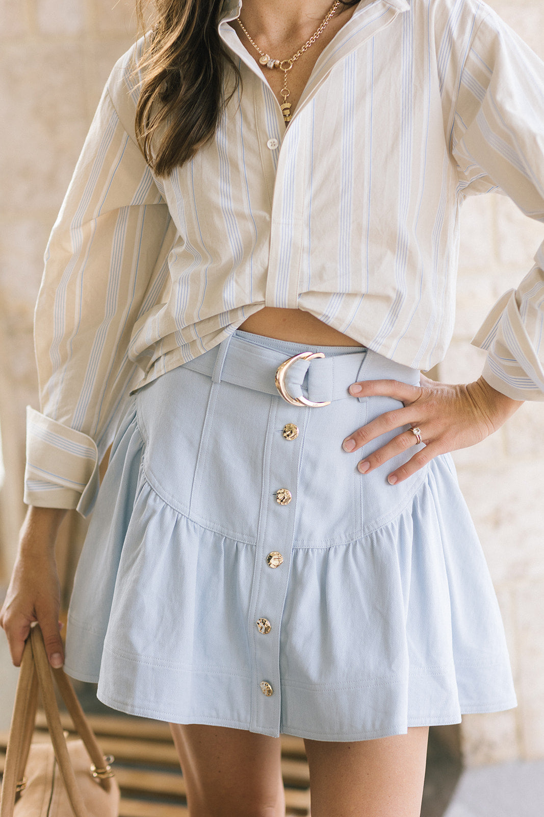 A woman wearing a cream and light blue striped button-up shirt and a light blue tiered mini skirt with gold-tone buttons and a crescent moon belt, holding a beige handbag.