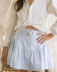 A woman wearing a cream and light blue striped button-up shirt and a light blue tiered mini skirt with gold-tone buttons and a crescent moon belt, holding a beige handbag.