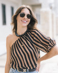 A woman wearing a black and tan striped asymmetrical one-shoulder blouse, sunglasses, and a beaded necklace, posing outdoors against a blurred light-colored building background.