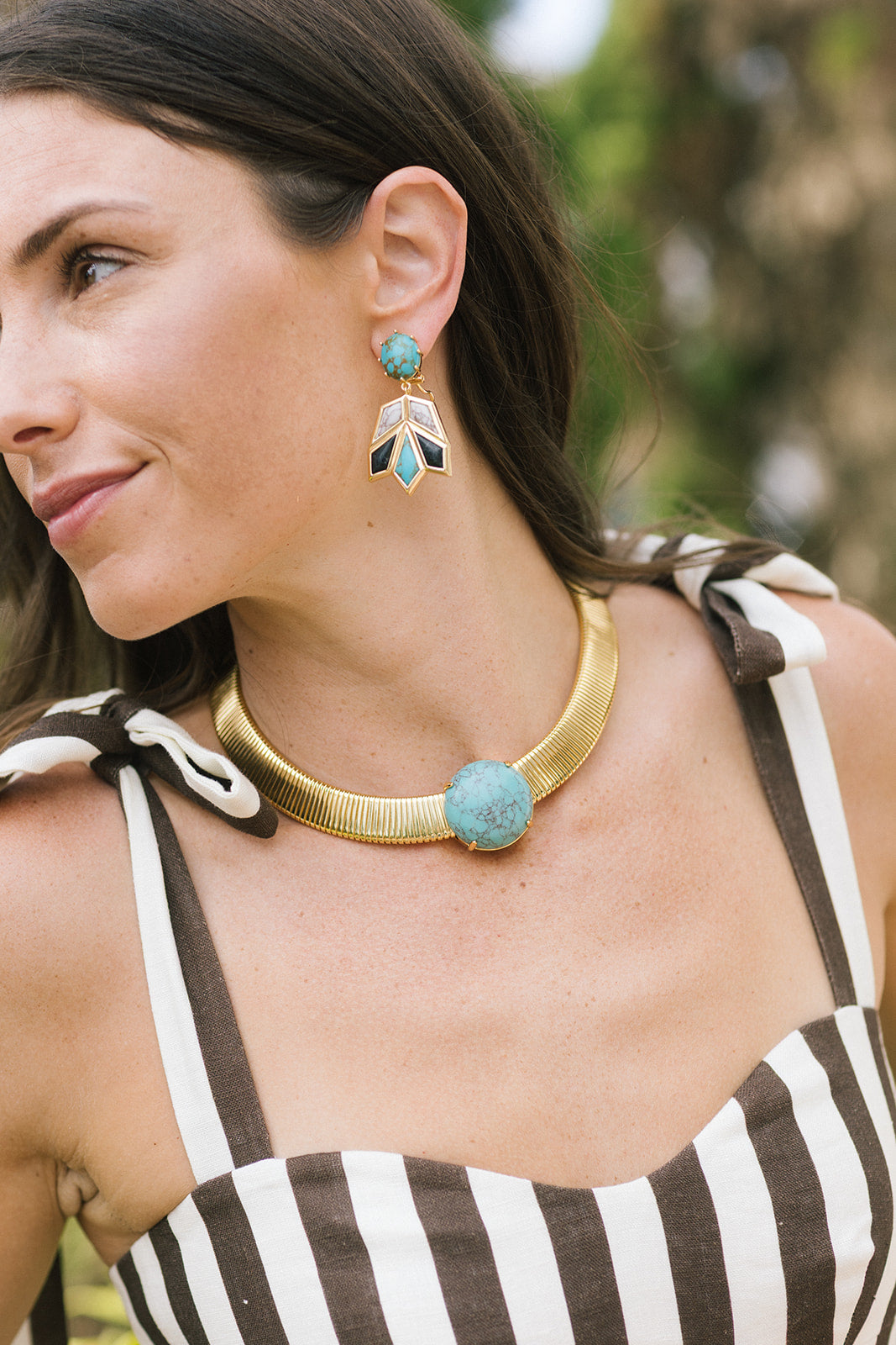 A close-up lifestyle shot of a woman wearing a gold-toned choker necklace with a round turquoise stone and matching drop earrings with turquoise, white, and gold feather-like details.