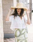 A woman wearing a white short-sleeve crop top and a white mini skirt with a green abstract pattern, accessorized with a wide-brimmed straw hat and a green choker, posing outdoors.