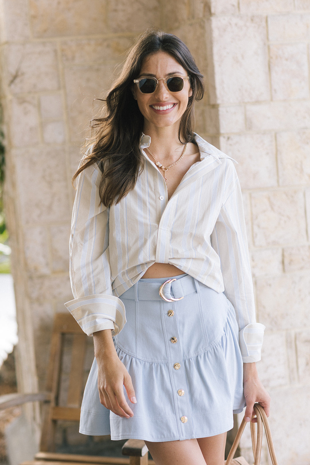A woman wearing a white striped button-down shirt and a light blue denim skirt with gold-tone buttons and a crescent moon detail at the waist, standing in front of a light stone wall.