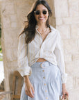 A woman wearing a white striped button-down shirt and a light blue denim skirt with gold-tone buttons and a crescent moon detail at the waist, standing in front of a light stone wall.