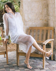 A woman wearing a sheer light blue long-sleeve set sits on a wooden bench in an outdoor patio setting with stone walls and tiled flooring.