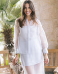 A woman wearing a sheer long-sleeve button-up blouse and matching skirt, holding a snakeskin-patterned clutch, standing outdoors in front of a stone wall and wooden bench.