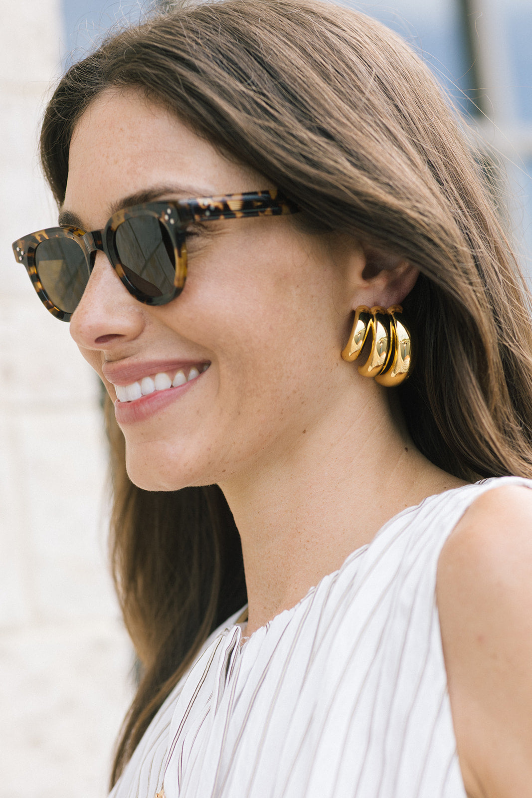 A close-up side profile of a woman wearing gold triple-hoop earrings and tortoiseshell sunglasses, with a white pleated top against a softly blurred light background.