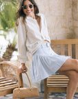 A woman wearing a cream long-sleeve button-up shirt and a light blue mini skirt sits on a wooden bench, holding a tan handbag.