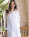 A woman wearing a light blue sheer long-sleeve button-up set, standing in front of a warm beige stone wall with soft green foliage in the background.