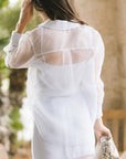 A woman viewed from behind wearing a sheer light blue long-sleeve shirt, standing in a softly blurred outdoor setting and holding a snakeskin-patterned clutch.