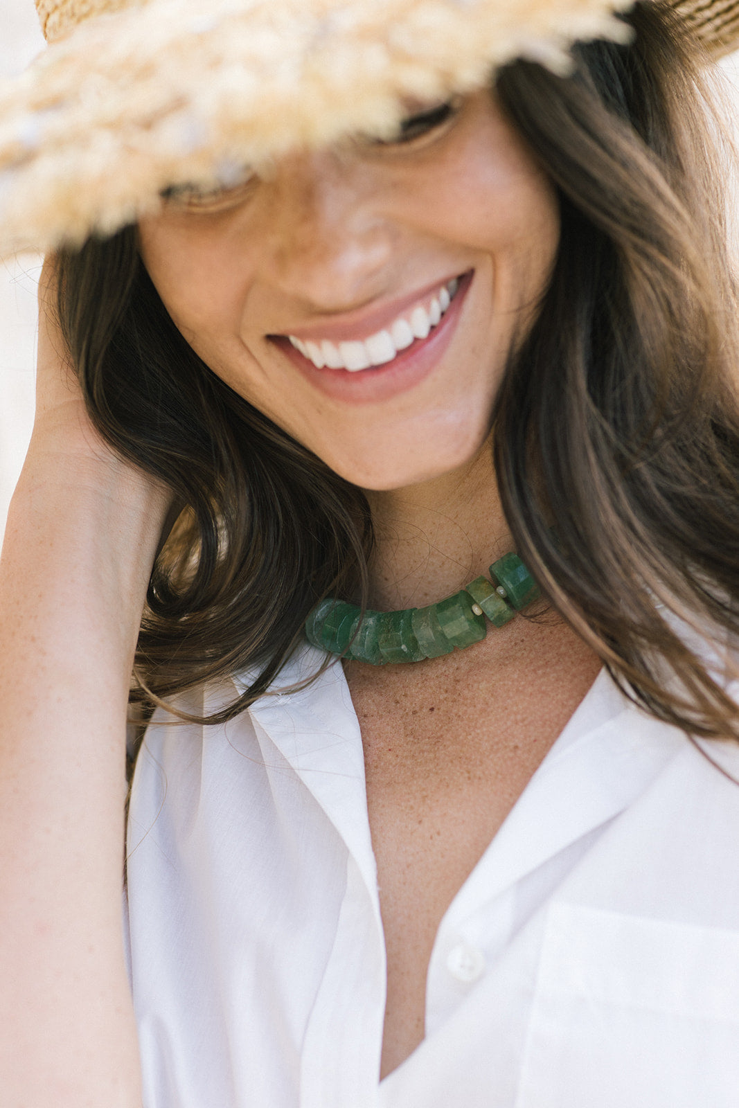 A close-up lifestyle shot of a woman wearing a chunky green stone beaded necklace, a white collared shirt, and a straw hat.