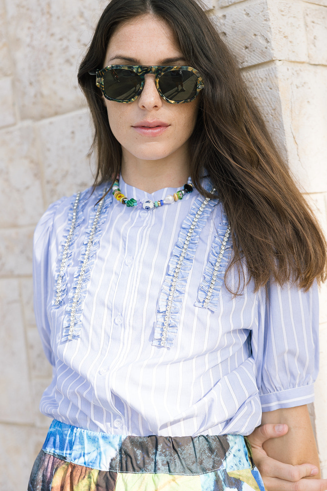 Woman wearing a light blue striped blouse with decorative elements, standing against a stone wall.