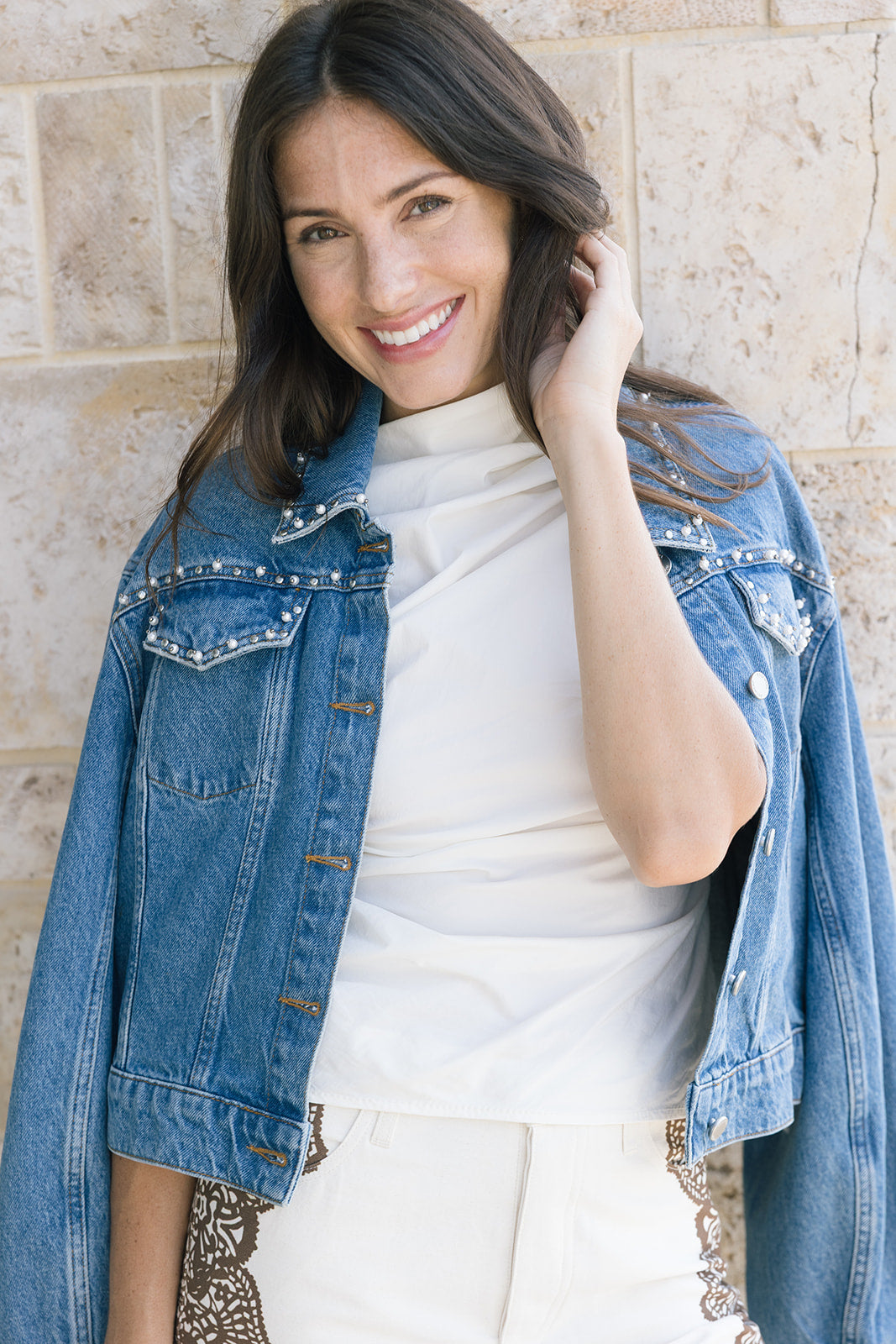 Woman wearing a denim jacket over a white top against a stone wall.