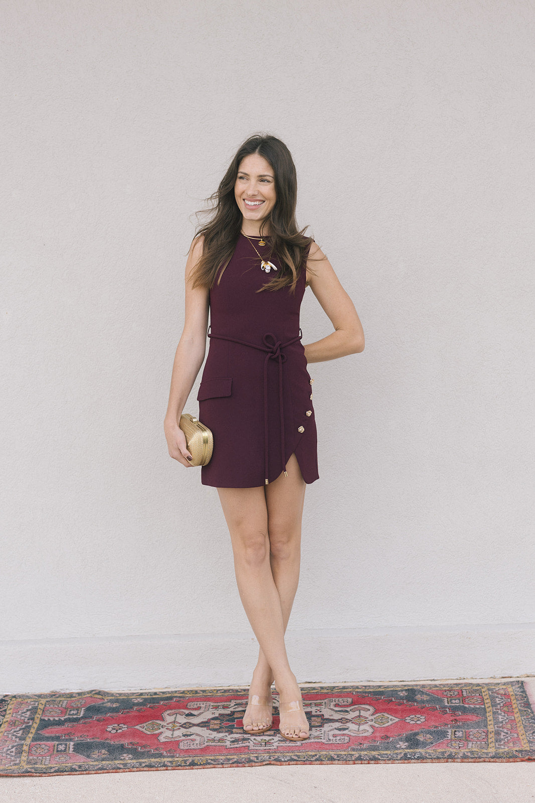 model wearing a merlot colored sleeveless mini dress holding a gold clutch on a grey background