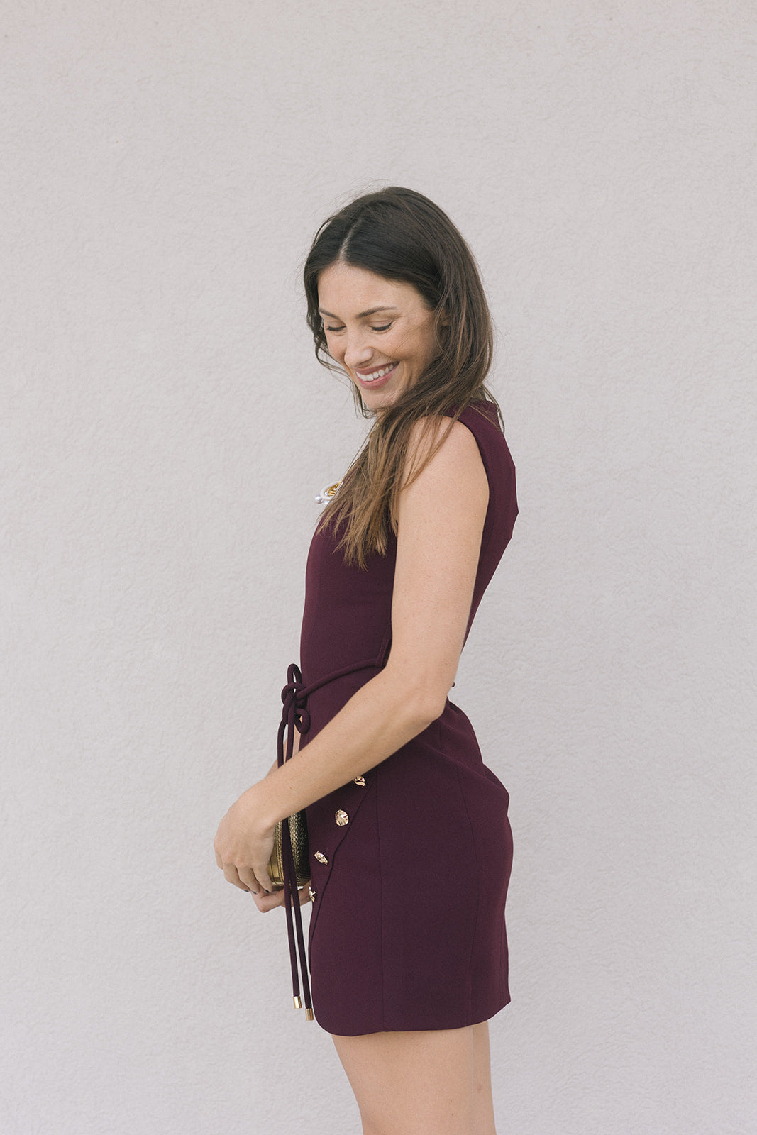 model wearing a merlot colored sleeveless mini dress holding a gold clutch on a grey background, side view