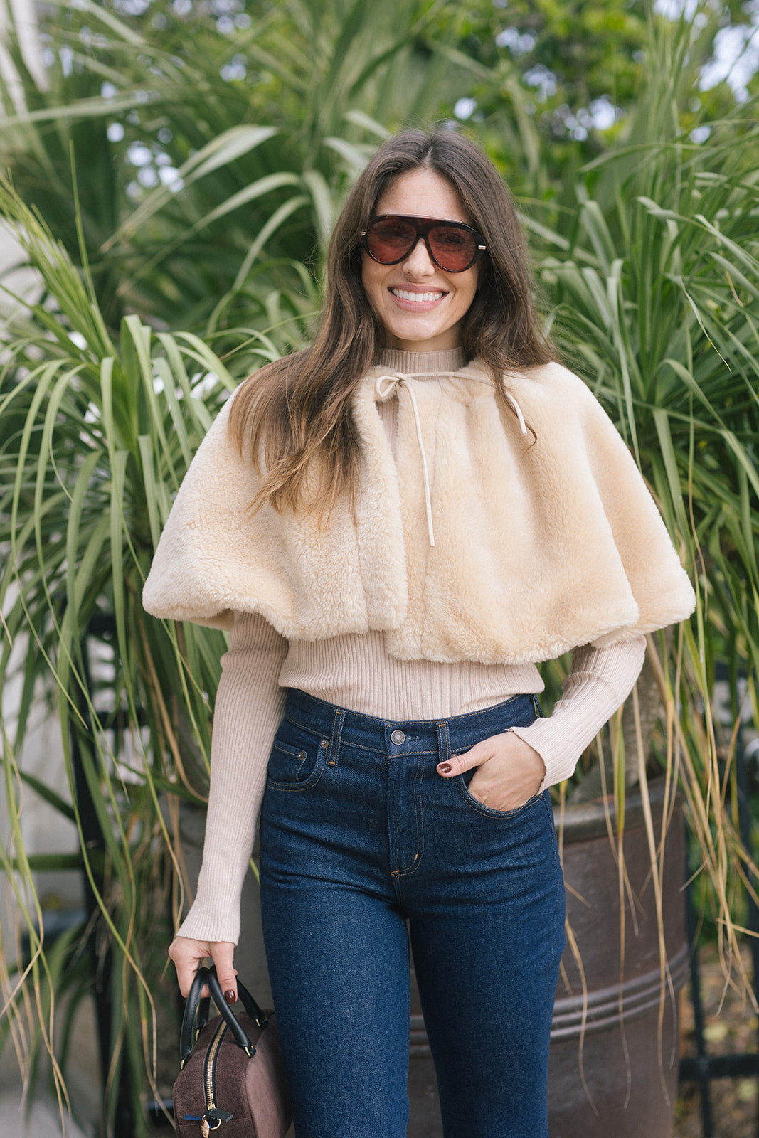 model wearing a tan fur cape over a long sleeve top with dark blue jeans outside 