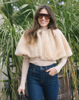 model wearing a tan fur cape over a long sleeve top with dark blue jeans outside 