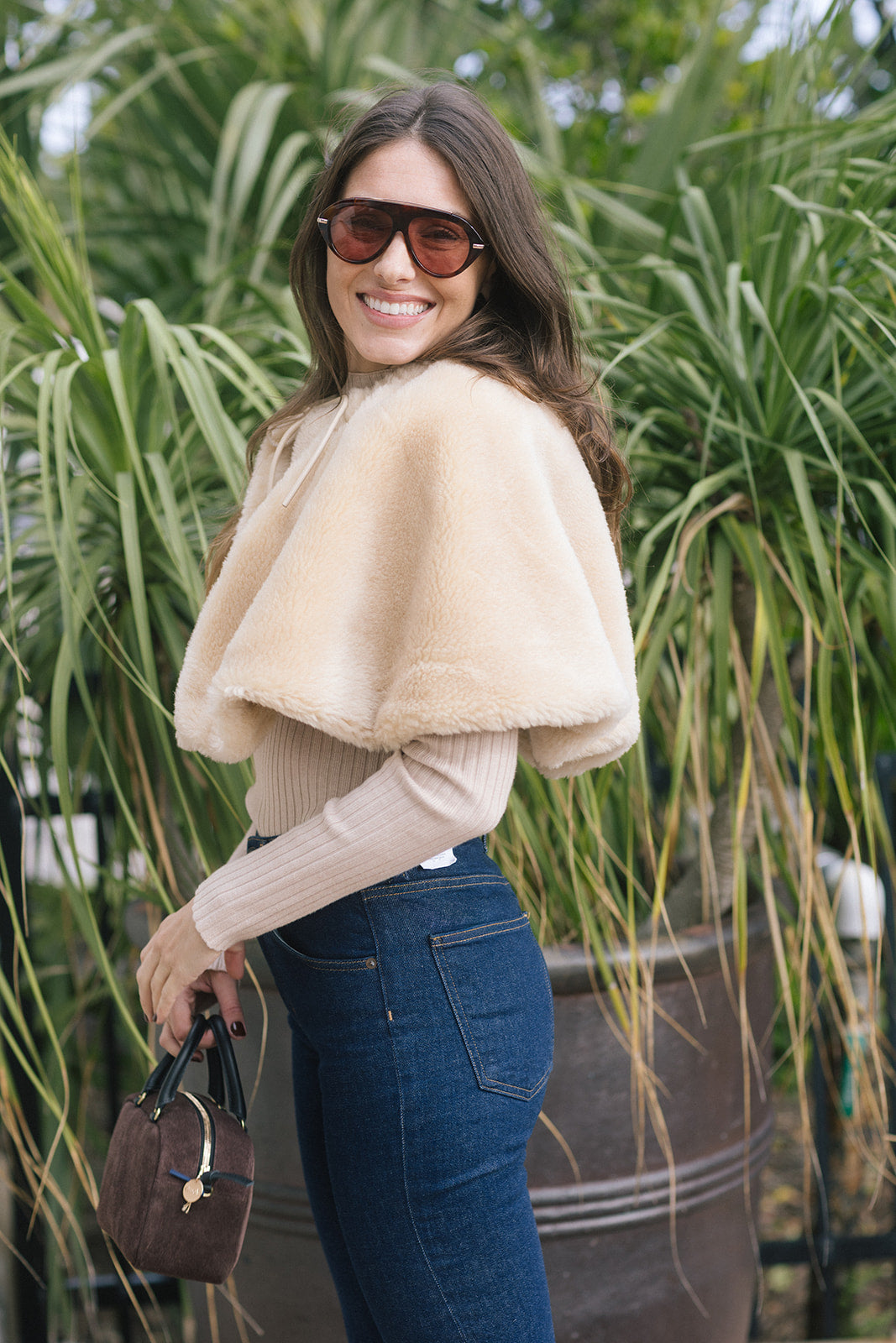 model wearing a tan fur cape over a long sleeve top with dark blue jeans outside, side view
