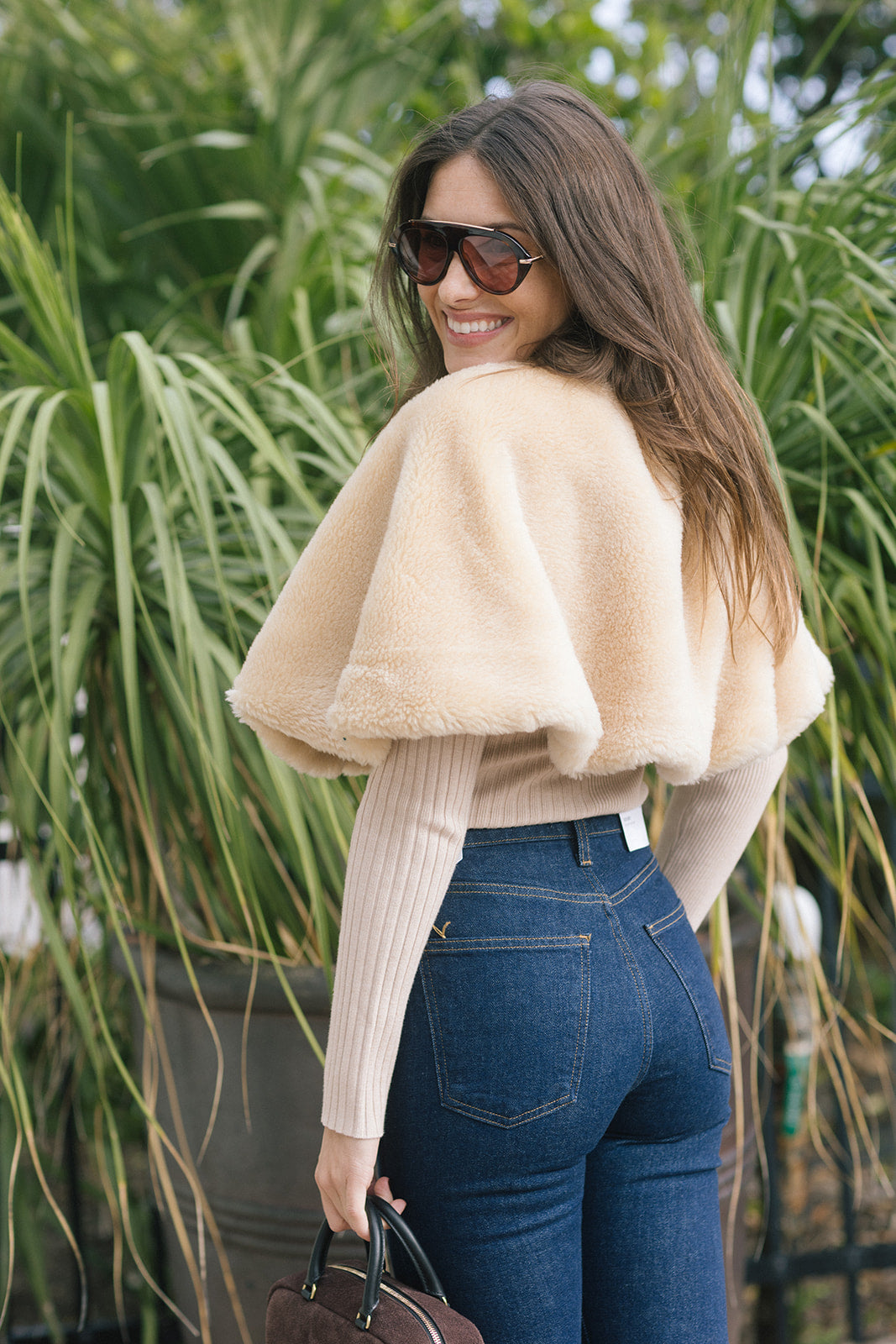 model wearing a tan fur cape over a long sleeve top with dark blue jeans outside, back view