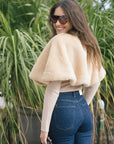 model wearing a tan fur cape over a long sleeve top with dark blue jeans outside, back view