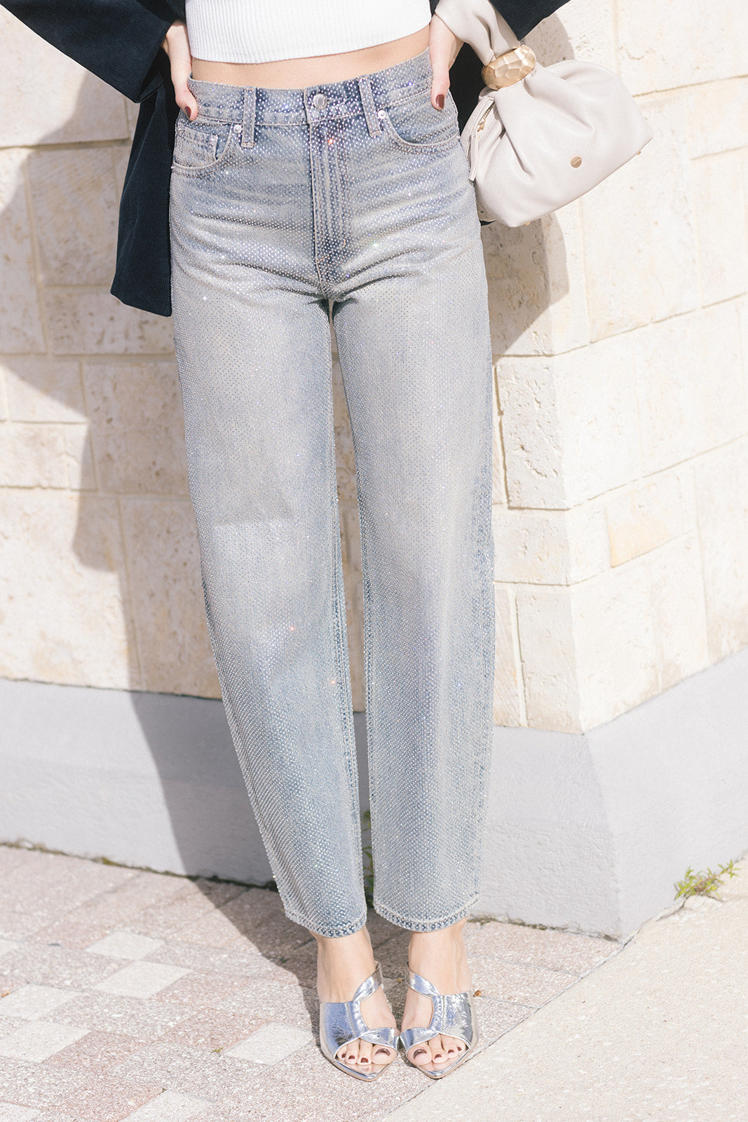model wearing light blue jeans covered in rhinestones and a white top with a dark jacket over holding a purse 