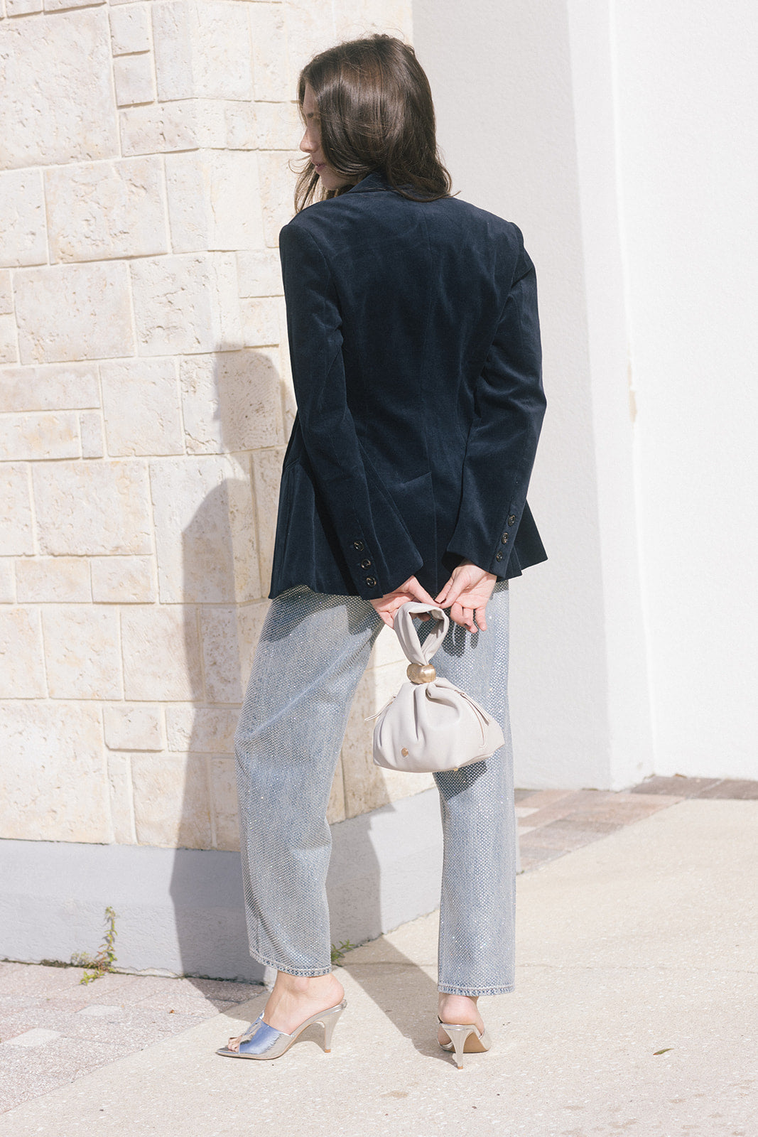 model wearing a navy blazer withe rhinestone jeans holding a cream bag, outside, back view