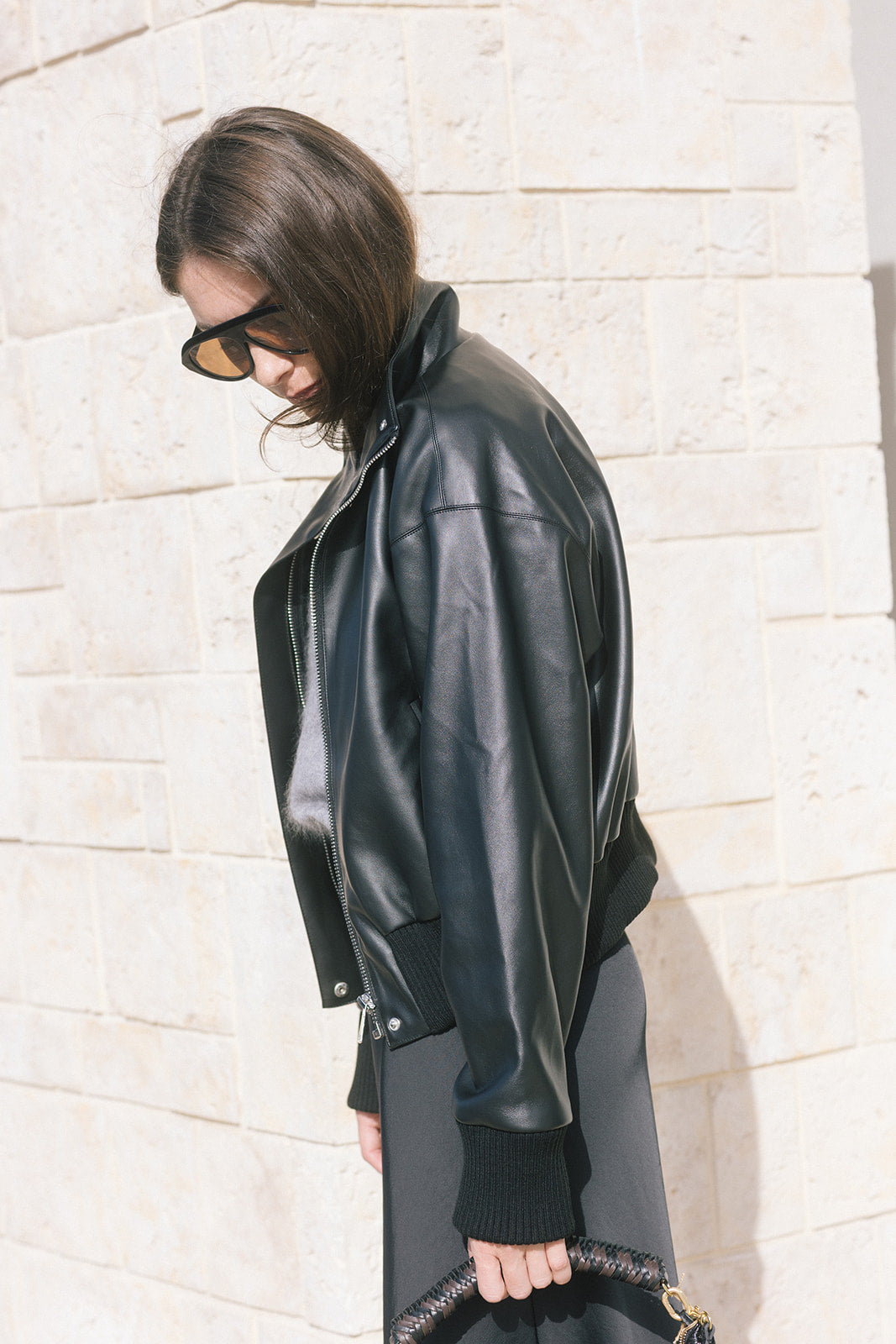 model wearing a black bomber jacket over a grey sweater with a black skirt while holding a black bag, side view