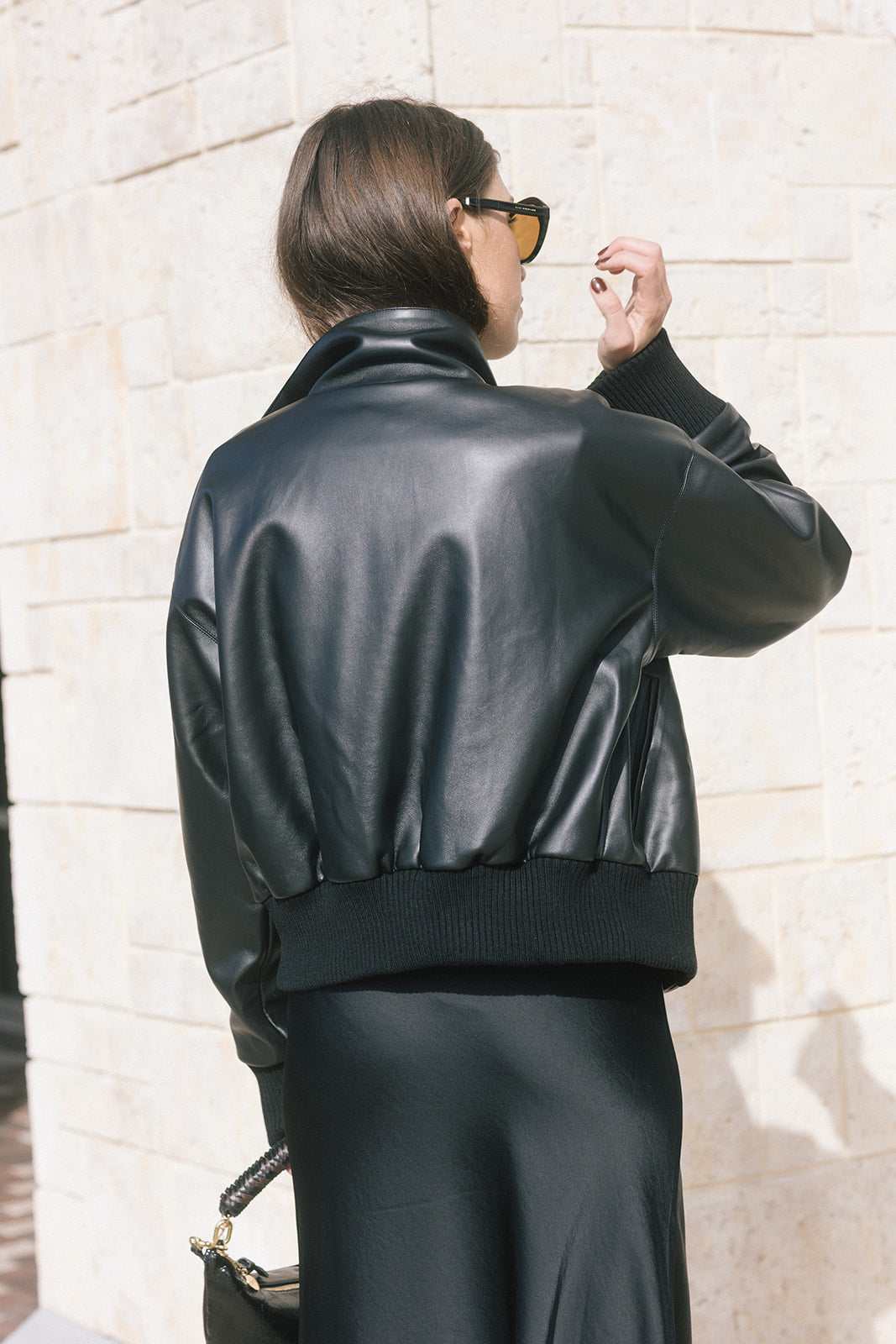model wearing a black bomber jacket over a grey sweater with a black skirt while holding a black bag, back view