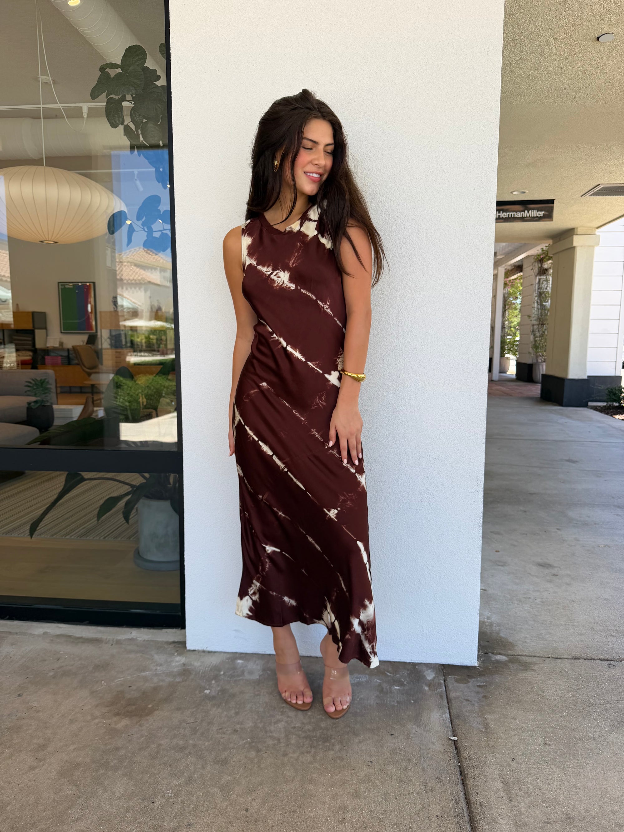 A woman wearing a sleeveless, high-neck, midi-length brown dress with a white tie-dye pattern, standing outdoors next to a white wall.