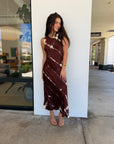 A woman wearing a sleeveless, high-neck, midi-length brown dress with a white tie-dye pattern, standing outdoors next to a white wall.