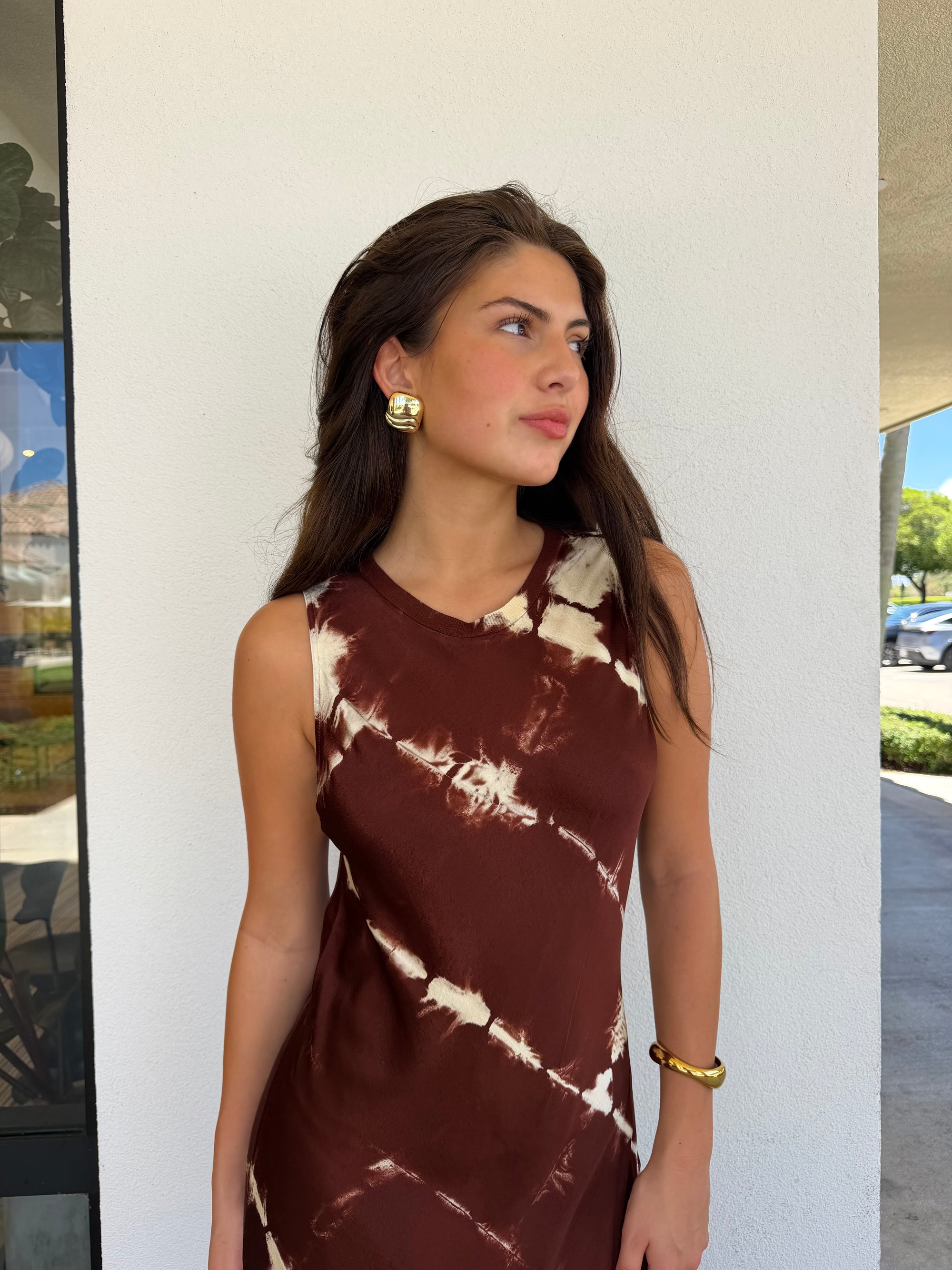 A woman wearing a sleeveless brown and cream tie-dye dress, standing outdoors against a white wall.