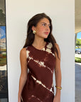 A woman wearing a sleeveless brown and cream tie-dye dress, standing outdoors against a white wall.