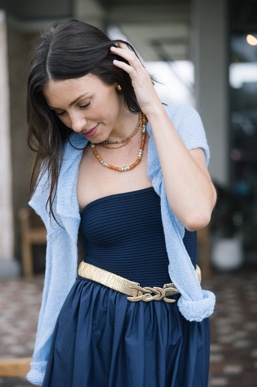Woman wearing By the Sea Necklace in Terracotta, a blue cardigan over a dark dress with a gold belt, indoors.