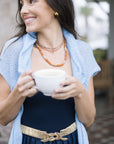 Woman holding a white mug outdoors, wearing By the Sea Necklace in Terracotta, a blue cardigan and dark dress with a gold belt.