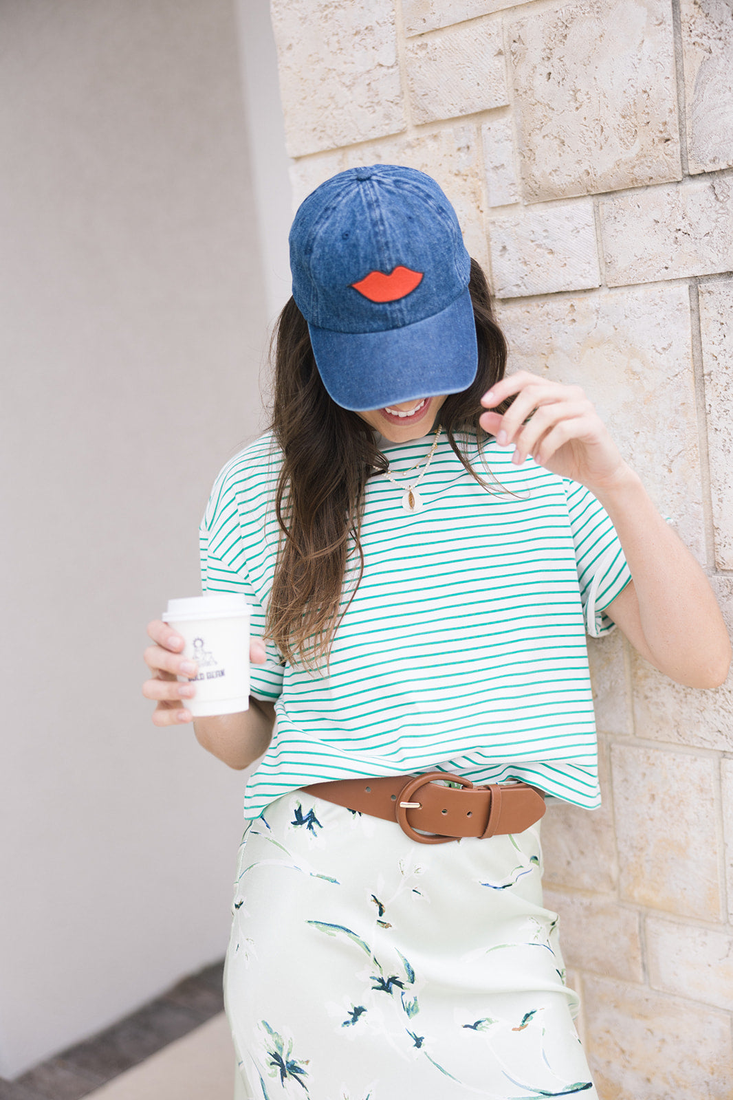 Woman wearing a blue cap, green striped shirt, and white skirt with floral pattern, holding a coffee cup.