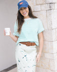 Woman wearing a striped shirt, cap, Kellen Belt, and Anya Print Skirt holding a coffee cup against a stone wall.