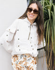 Woman wearing a white jacket with floral shorts standing next to a plant.