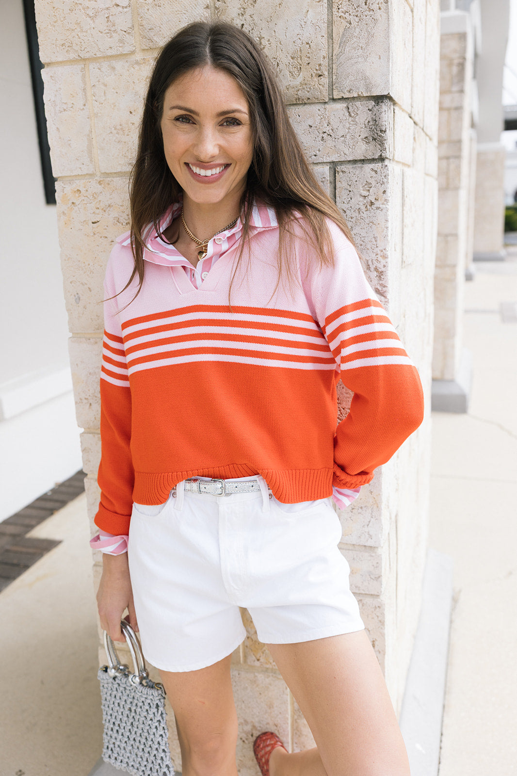 Woman wearing a pink and orange striped sweater with white shorts, standing against a stone wall.