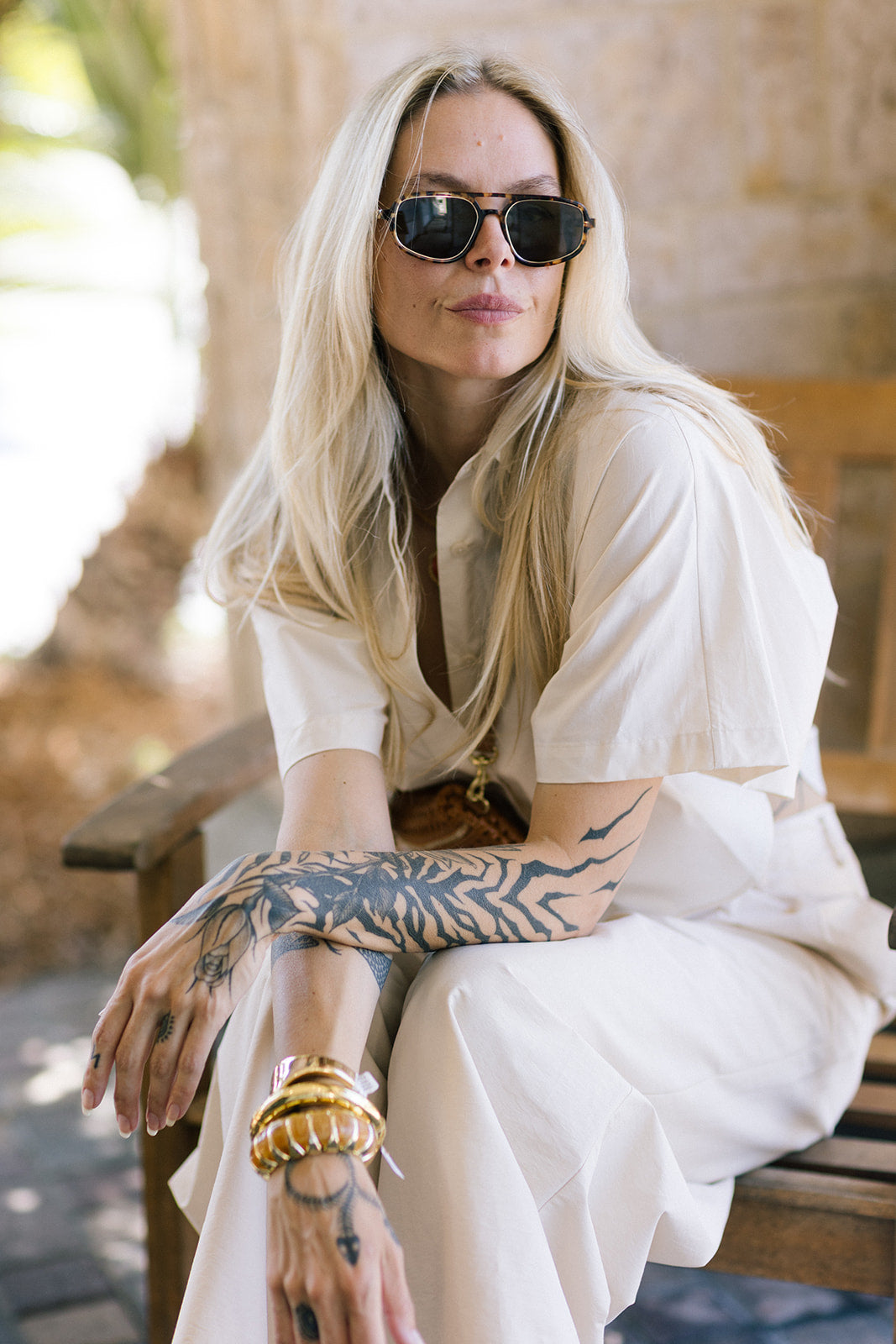 Woman with tattoos wearing sunglasses and a white outfit, sitting outdoors.
