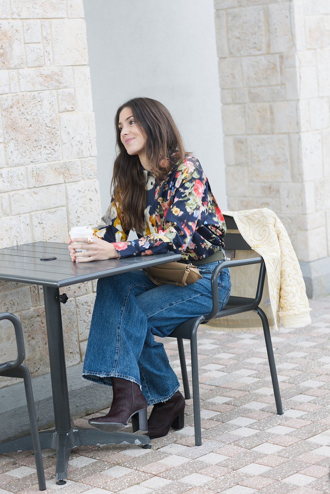 Woman sitting at an outdoor table with a cup, wearing a floral top and jeans.