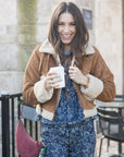 Woman holding a coffee cup and Lune Bag in Currant outdoors, wearing a brown jacket with shearling lining and blue floral outfit.