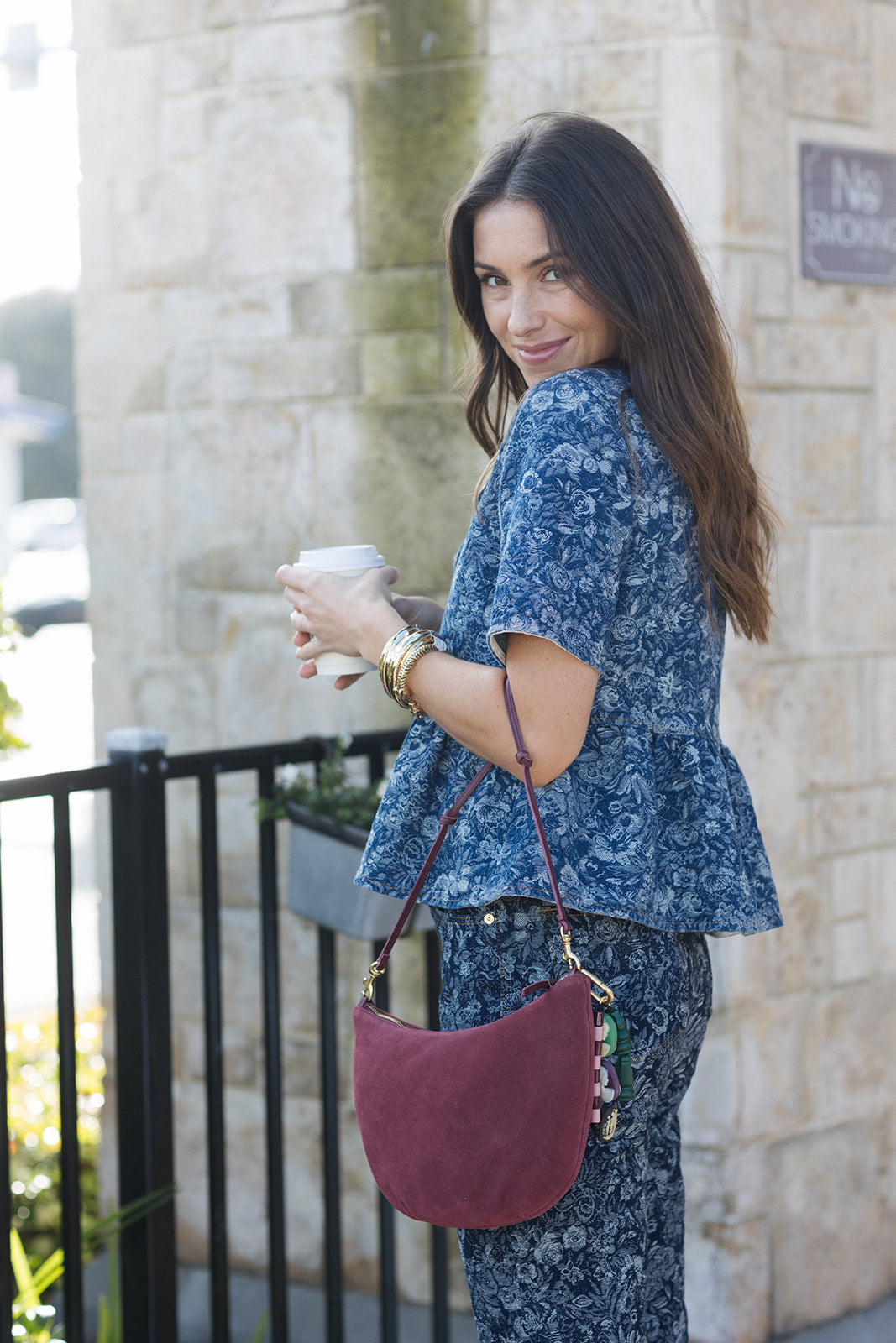 Woman holding a coffee cup with a maroon handbag, standing against a stone wall.