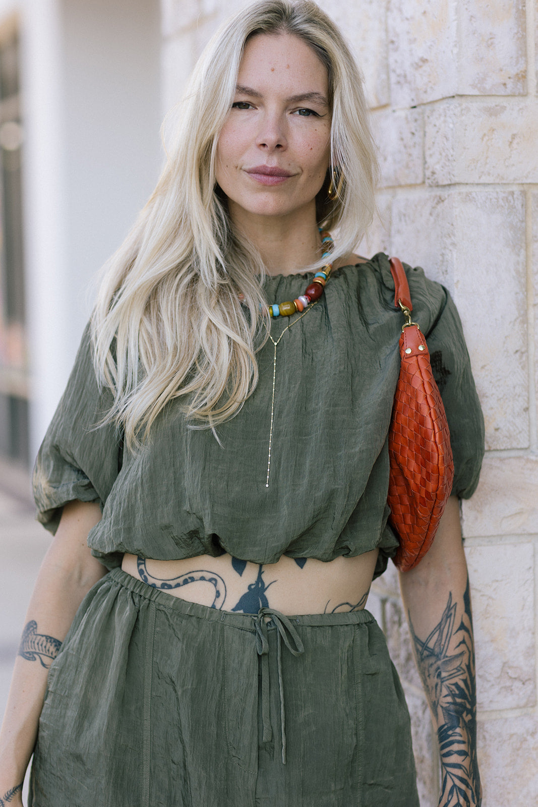 Woman wearing an olive green outfit with a red bag against a stone wall.