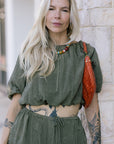 Woman wearing an olive green outfit with a red bag against a stone wall.