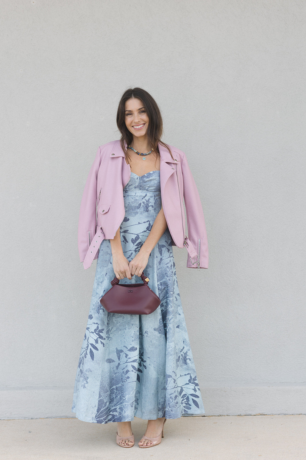 Woman wearing a pink jacket over a floral dress with a maroon handbag against a plain background