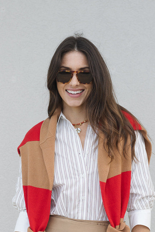 Woman wearing a red and beige color-blocked cardigan over a striped shirt with Zander Hawksbill Sunglasses against a gray background