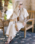 Woman in a white outfit sitting on a wooden chair outdoors.