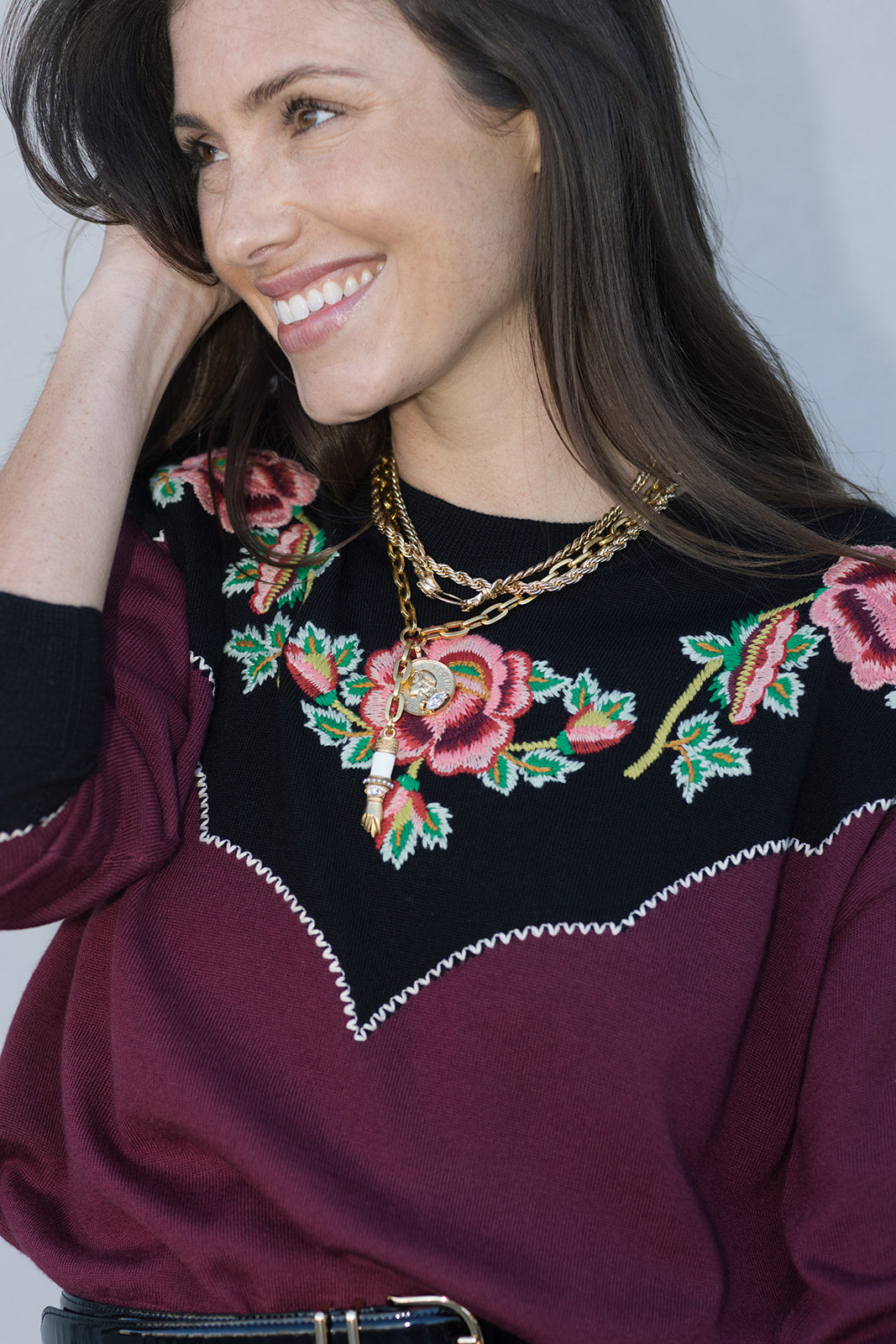 Woman wearing a burgundy sweater with floral embroidery and gold jewelry.