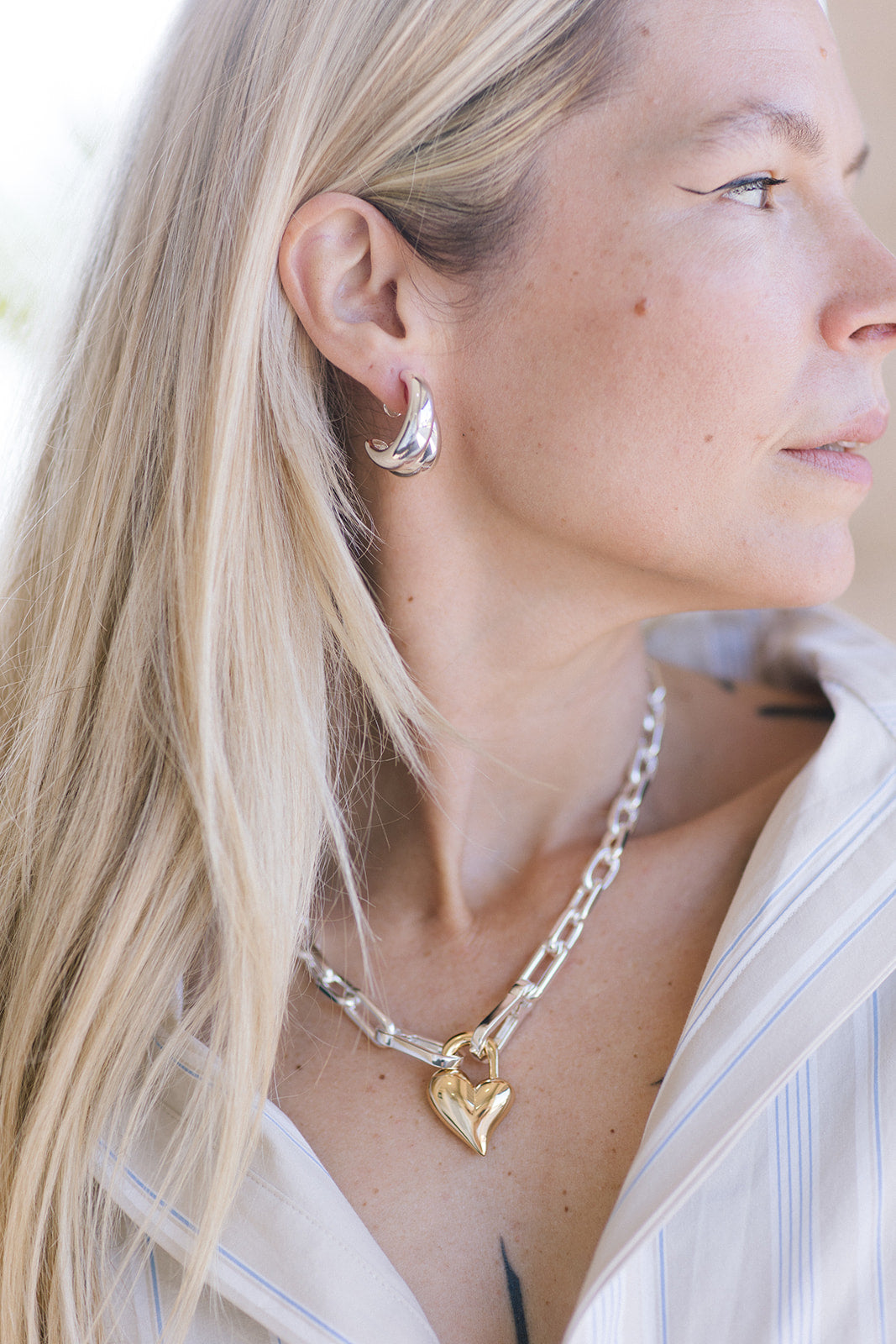 Close-up of a person wearing a gold heart-shaped necklace and silver earrings.