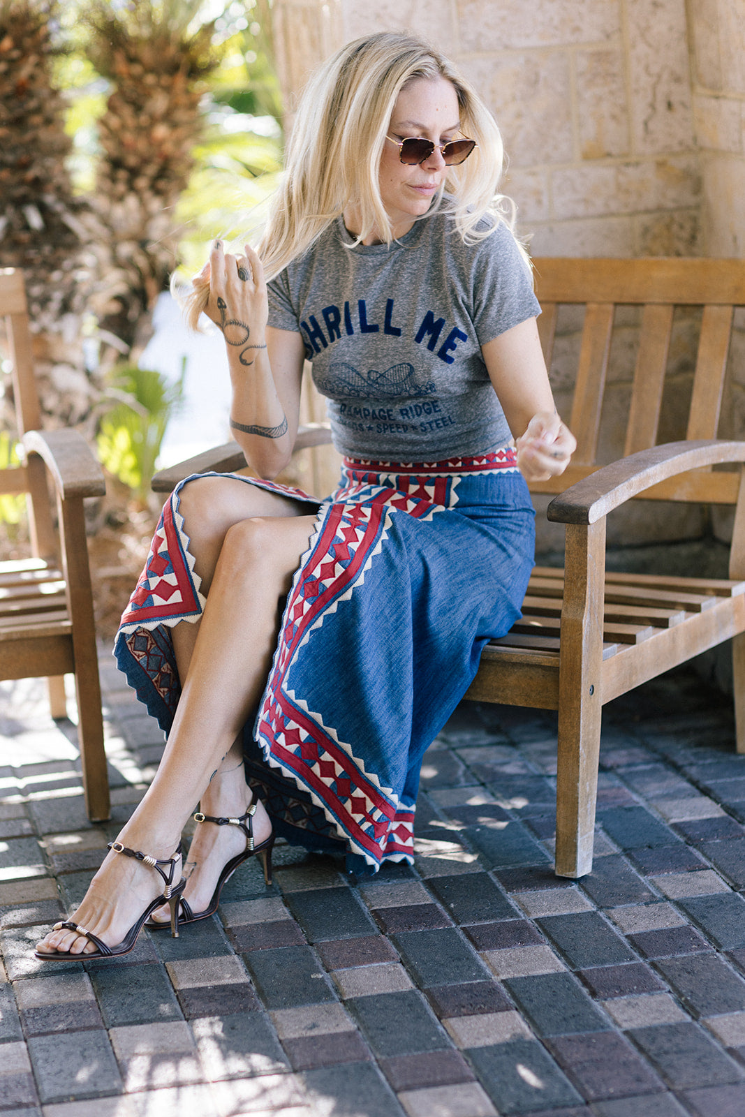 Woman sitting on a wooden chair wearing a gray t-shirt and blue patterned skirt.