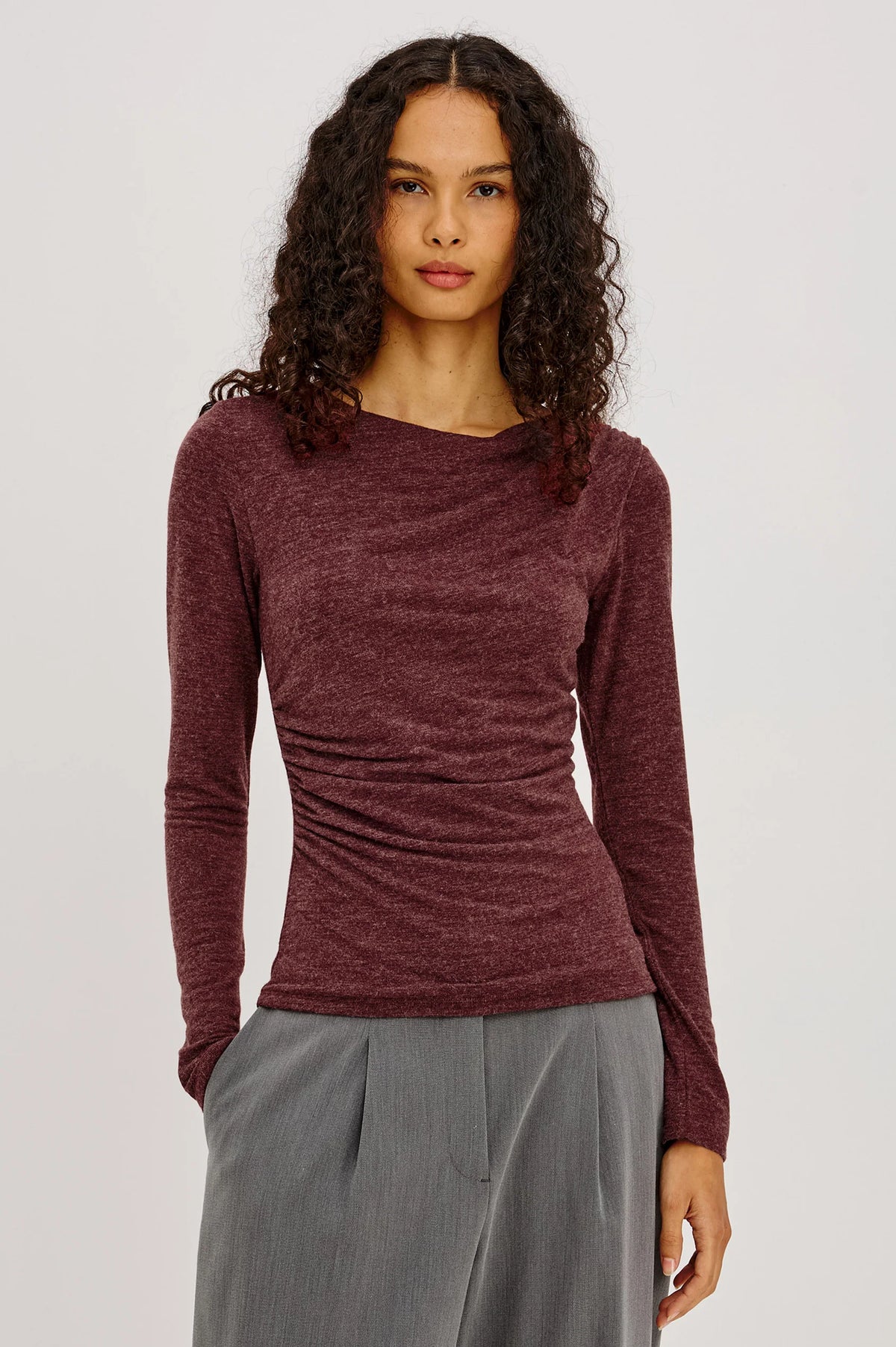 model wearing a long sleeve merlot top with a cowl neck and grey pants on a light grey background 